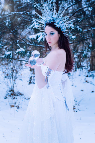 Fantasy photograph of a woman in a white glittery gown and elaborate headdress of icicles and silver glittered flowers, holding a pyramid of clear crystal spheres in the snowy forest.