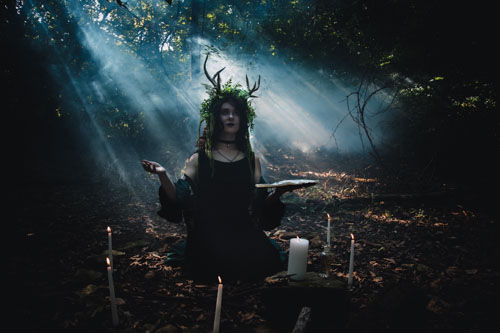 Fantasy photograph of a non-binary person in a black dress and headdress of leaves, vines, and antlers, kneeling in the forest surrounded by candles, holding a spellbook, with the sunlight streaming through the fog behind. Kansas City