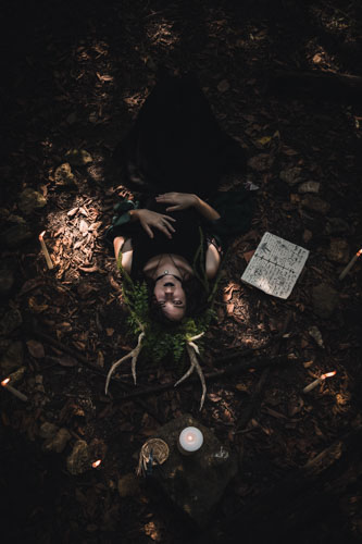 Fantasy photograph of a non-binary person in a black dress and headdress of leaves, vines, and antlers, lying on the ground upside down looking up at the camera, surrounded by candles, stones, and spellbooks. - Kansas City