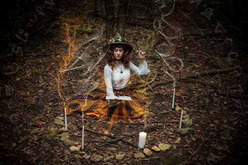 Fantasy photograph of a woman in an orange skirt, white lace shirt, and brown witch hat holding a glowing spellbook and appearing to use her hands to cast a spell, surrounded by a circle of stones and candles in the woods. Colored smoke rises from the candles, and arcane symbols encircle her.